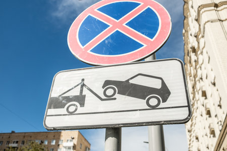 Bright no parking sign with towing graphic prominently displayed above vehicles being towed in a bustling city environment under clear skies.の写真素材
