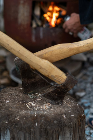 A person uses a sharp ax on a log, preparing firewood while a fire crackles in the background,...