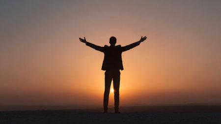 A man stands confidently with arms raised in celebration at sunset, showing the warm colors of the sky and the tranquil atmosphere around him.の素材