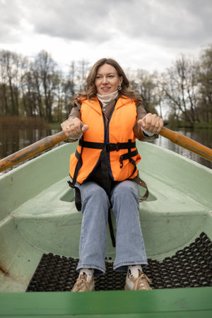 A woman wearing an orange life vest rows a green boat on a quiet lake, with trees lining the shore under a cloudy sky.の写真素材