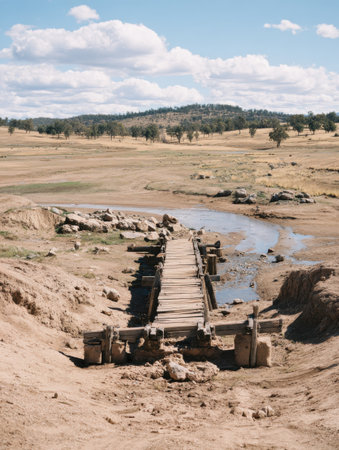 A wooden bridge stands over a parched riverbed in a vast countryside. A few clouds float above a gentle hill, creating a serene backdrop.の素材