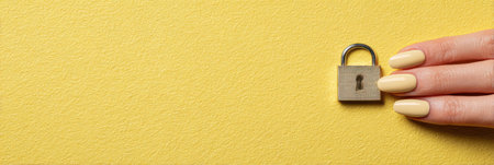 A hand with neatly manicured nails holds a small metallic lock against a vibrant yellow backdrop, emphasizing a sense of security and simplicity.の素材