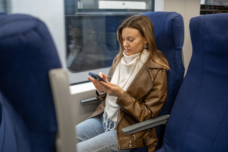 A young woman in a brown leather jacket sits on a train, absorbed in her smartphone, showcasing a...