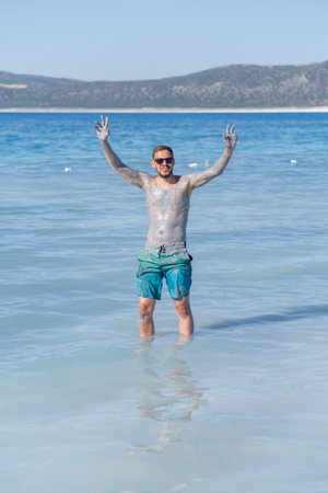 Person stands in shallow water with mud covering his body while smiling and raising his hands in joy at a beautiful beach under a clear sky.の写真素材