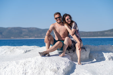 Two people relax on white rocks near the sea, smiling and enjoying a sunny day at a beach, with mountains in the background and clear blue skies.の写真素材