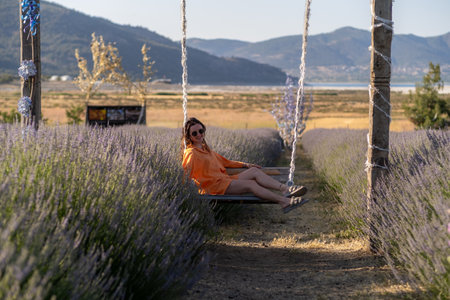 Bright sunlight illuminates a woman lounging on a swing surrounded by vibrant lavender plants in a peaceful rural area.の写真素材