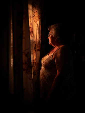 An elderly woman by the curtain gazes out the window, warm evening light illuminating her face in a serene, nostalgic moment.の素材