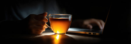 A hand holds a glass cup of warm tea while another hand types on a laptop in a dimly lit setting during the evening, creating a calm atmosphere.の素材