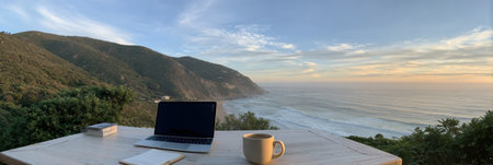 Enjoying a peaceful morning at a coastal workspace with a laptop and coffee, while waves crash against the shore and mountains rise in the background.の素材