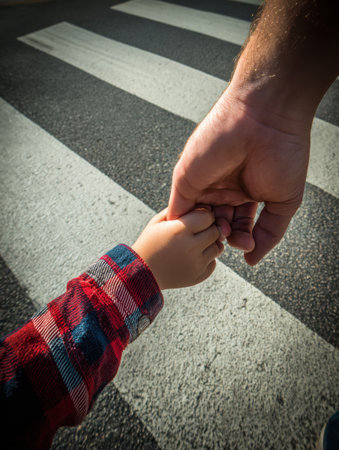 Hands of a child and an adult are seen holding each other while crossing a zebra crossing, symbolizing safety and love during a sunny day.の素材