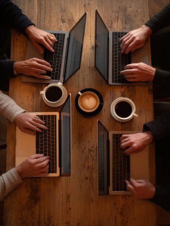 Four hands typing on laptops are seen around a wooden table, with coffee cups nearby, creating a relaxed setting perfect for productivity.の素材