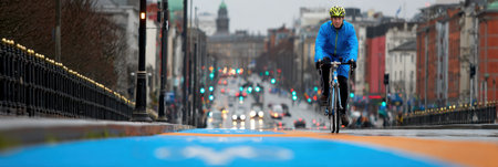 A cyclist wearing a blue rain jacket navigates a bright bike lane amid a bustling urban setting on a rainy evening with traffic lights glowing.の素材