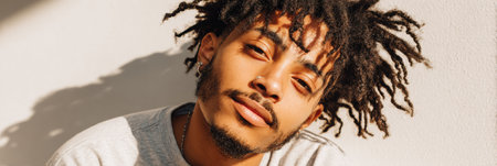 A young man with dreadlocks leans against a wall, bathed in warm evening light, capturing a moment of calm and confidence in a trendy outfit.の素材