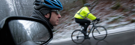 A cyclist is seen riding along a snow-covered road while a driver watches closely through the side mirror, highlighting the cold winter scene.の素材