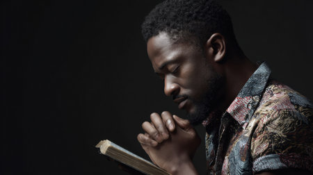 A man holds a book close, eyes closed, and fingers clasped as he reflects quietly in a dimly lit space, immersed in contemplation.の素材