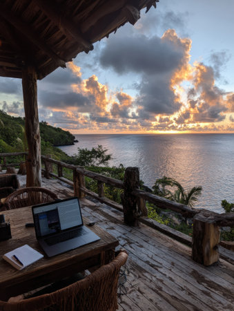 A peaceful workspace features a laptop on a wooden table, with a beautiful sunset and calm ocean waves in the background, creating an inspiring atmosphere.の素材