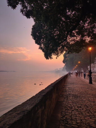 People take leisurely walks by the riverbank as the sun sets, painting the sky with warm colors while trees line the picturesque pathway.の素材