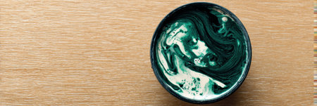 A circular bowl contains rich green paint with swirling patterns, placed on a smooth wooden table. Soft natural light enhances the colors.の素材