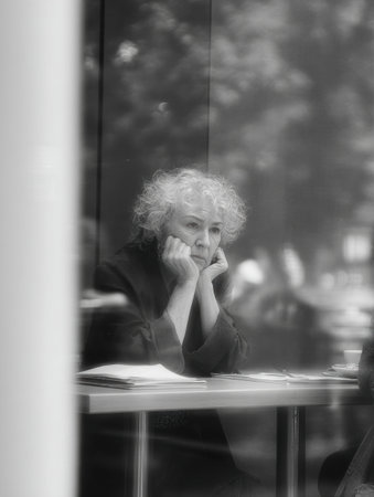 A woman with curly hair rests her chin on her hands, lost in thought, as she looks out through a glass window on a calm day in the city.の素材