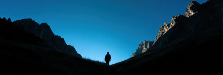 A solitary hiker walks along a path in a mountainous area during dusk. The sky is clear and blue, with towering cliffs on either side.の素材