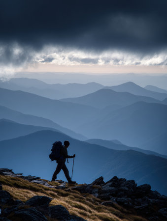 A solitary hiker navigates a rocky mountain ridge surrounded by layers of distant peaks under a moody sky at twilight.の素材