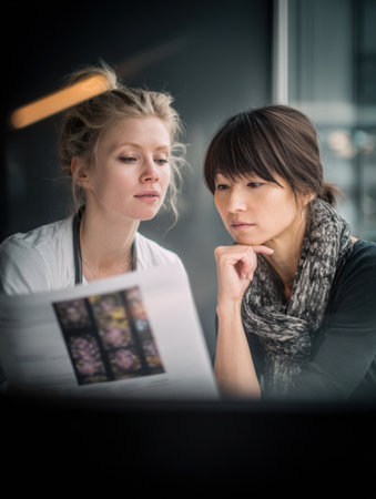Two women are engaged in a focused conversation about a design project while seated in a stylish cafe with large windows letting in natural light.の素材