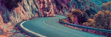 A winding road leads through a mountainous area, flanked by lush trees showcasing vibrant colors under bright daylight. Scenic view invites exploration.の素材