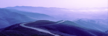 A peaceful landscape showing gentle hills and a winding road at dawn. The soft purple light creates a serene and tranquil atmosphere filled with mist.の素材