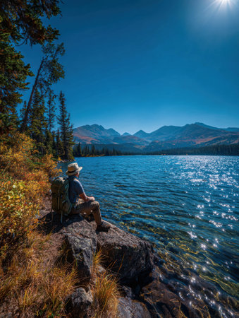 A person sits on a rock by a sparkling lake surrounded by mountains and trees, soaking in the peaceful atmosphere during the warm afternoon hours.の素材