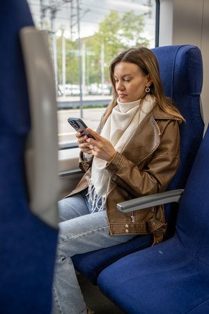 Young woman with long hair and a brown jacket is focused on her smartphone while relaxing in a train...