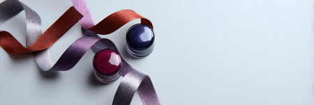Burgundy and navy nail polish containers are next to several shiny ribbons on a plain surface.の素材