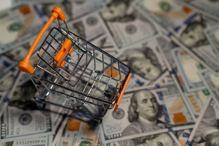 A small shopping cart is set on a pile of hundred dollar bills, highlighting the connection between shopping and money.の写真素材