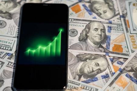 A smartphone shows a rising graph symbolizing financial growth, placed on a backdrop of US dollar bills.の写真素材
