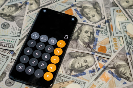 A calculator is placed on top of numerous one hundred dollar bills, symbolizing financial calculations and management.の写真素材