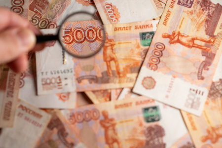 A hand holds a magnifying glass over a 5000 ruble note, surrounded by various banknotes in a...