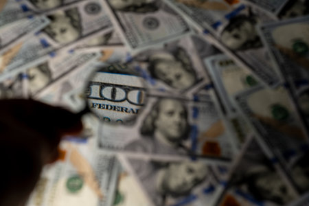A hand uses a magnifying glass to inspect a one hundred dollar bill amid a spread of cash on a table.の写真素材