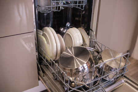 Clean plates and stainless steel pots are neatly arranged in a dishwasher in a contemporary kitchen setting.の写真素材