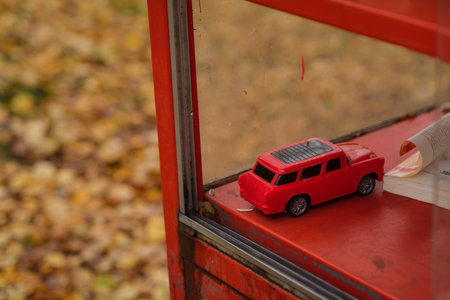 A red toy car sits quietly on a ledge of a phone booth surrounded by colorful autumn leaves in a...