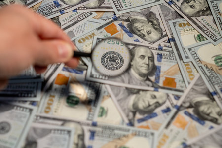 A hand holds a magnifying glass over several hundred dollar bills, focusing on the intricate details of US currency.の写真素材