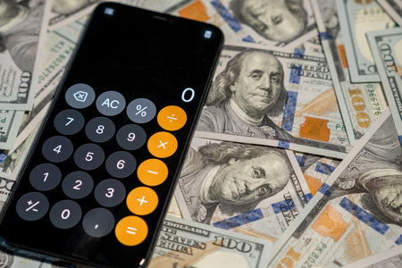 A smartphone with a calculator rests on a pile of hundred-dollar bills, emphasizing budget and finance management.の写真素材