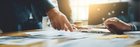 Two professionals engage in a serious business discussion while pointing at documents and plans on a wooden table in an office.の素材