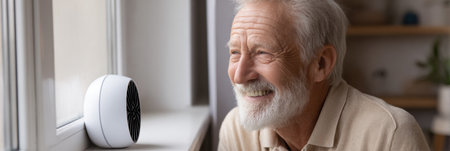 A senior man sits comfortably by the window, smiling as he enjoys the sunlight and a gentle breeze from a nearby fan in his cozy space.の素材