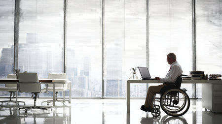 A man in a wheelchair focuses on his laptop in a spacious, light-filled office with a view of the cityscape beyond.の素材
