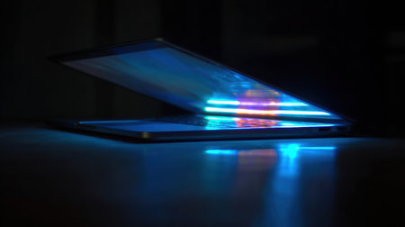 Bright blue and red lights illuminate the laptop as it reflects on a smooth surface in a dimly lit setting at night.の素材