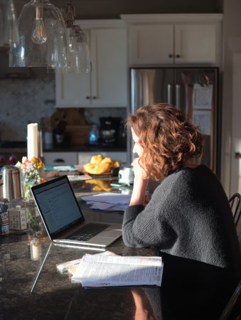 Focused individual engages with a laptop at a kitchen counter surrounded by morning light and fruit, immersed in work or study.の素材