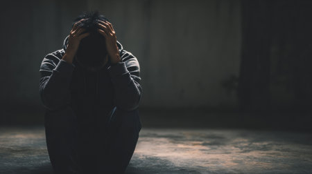 A man sits on the floor with his hands on his head, reflecting feelings of emotional stress and depression in a dimly lit environment.の素材