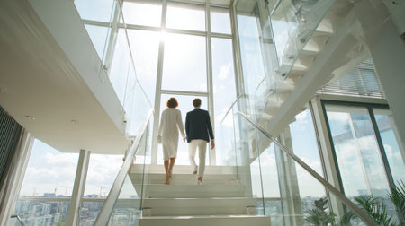 Young couple walks hand in hand up the stairs of a stylish apartment filled with natural light and city views.の素材