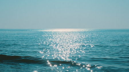 Bright sunlight dances on the calm ocean, creating a peaceful atmosphere as waves gently lap against the shore during midday.の素材