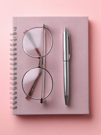 A flat lay display features stylish glasses, a sleek pen, and a simple notebook arranged on a soft pastel pink surface, ideal for a workspace.の素材