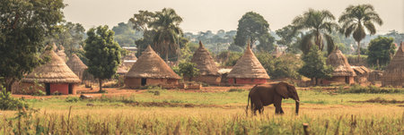 An African elephant roams through a grassy field near a village characterized by traditional huts, highlighting the cultural heritage of the area.の素材